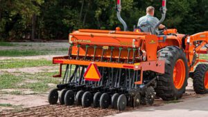 Person driving a tractor tilling land with a Land Pride 3P606NT Compact Drill attachment on the back.