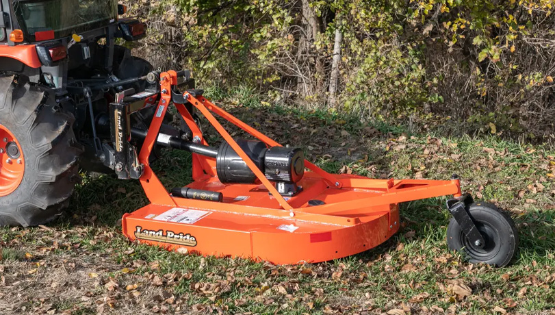Land Pride RCR15 Series Rotary Cutter attachment attached to the back of a tractor in a field with brushes and trees in the background.