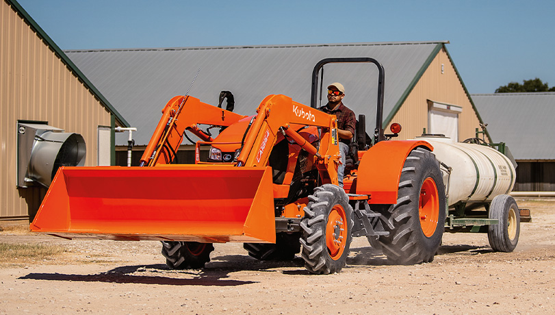 New Kubota M5-111HD12 Tractor towing a water tank with storage buildings in the background.