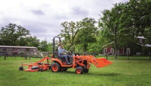 Person driving a Kubota tractor with excavator and rotary cutter attachment in a field.