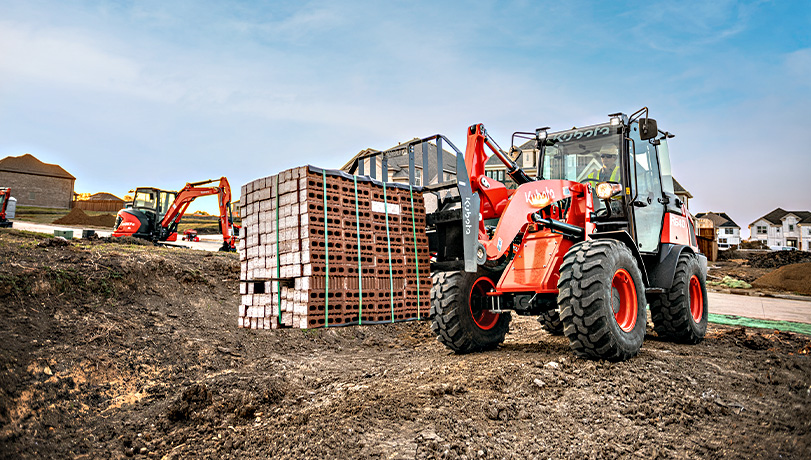 Person on a construction site driving a Kubota R640 Wheel Loader carrying a stack of bricks.