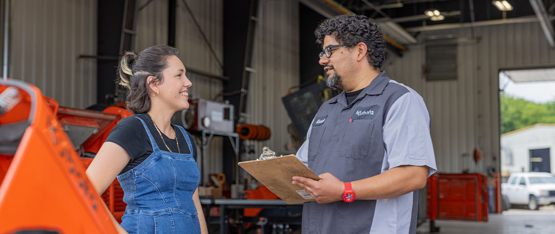 A service advisor and a customer discussing various options for their Kubota or Hitachi product in a mechanic bay.