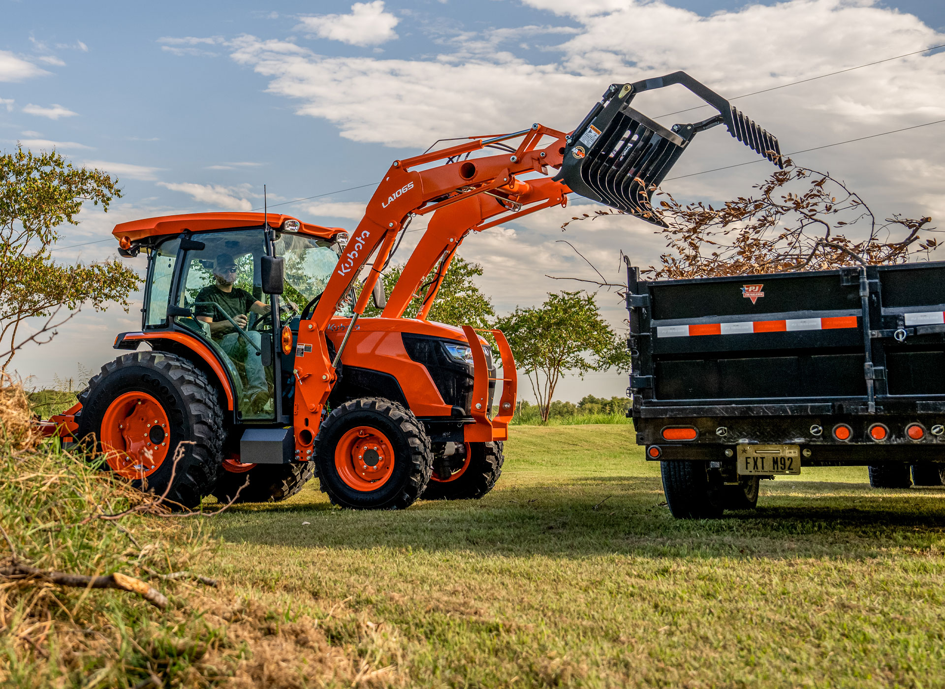 Person in a Kubota tractor with a skeleton grapple lifting tree branches into a truck with a dump bed.