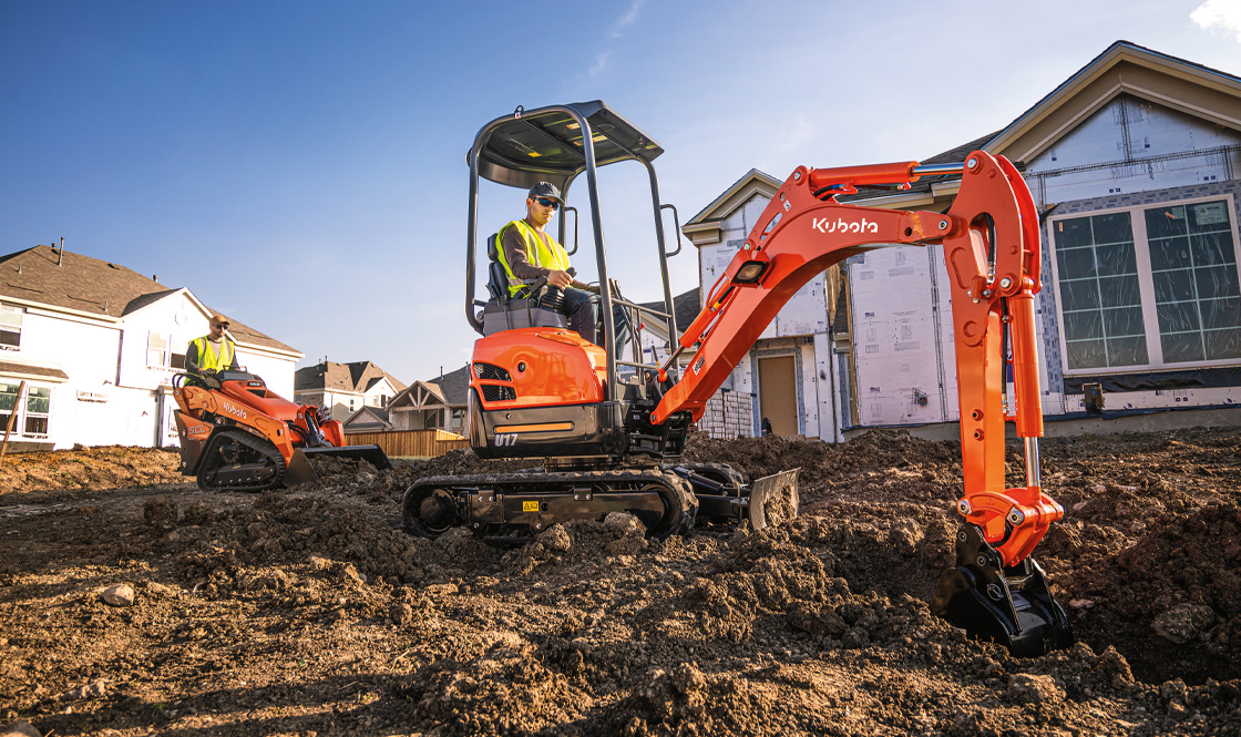 Woman on a tractor with a few different attachments on it driving on a gravel road during a sunny day.