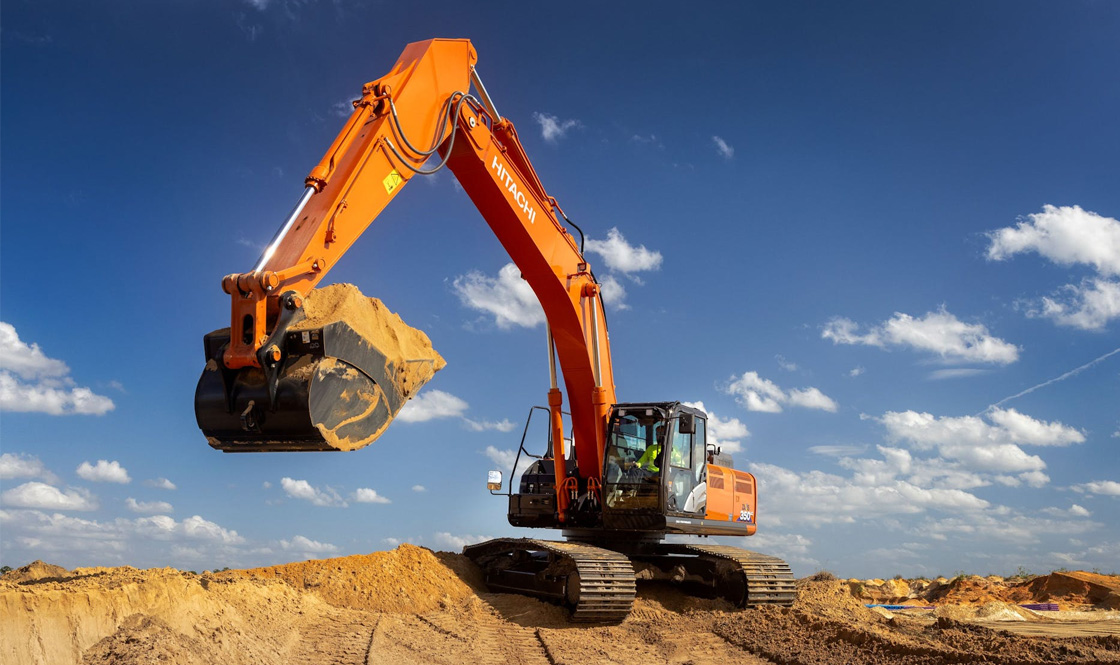 Person in a large orange Hitachi excavator clearing land on a construction site on a clear day with rugged terrain in the background.