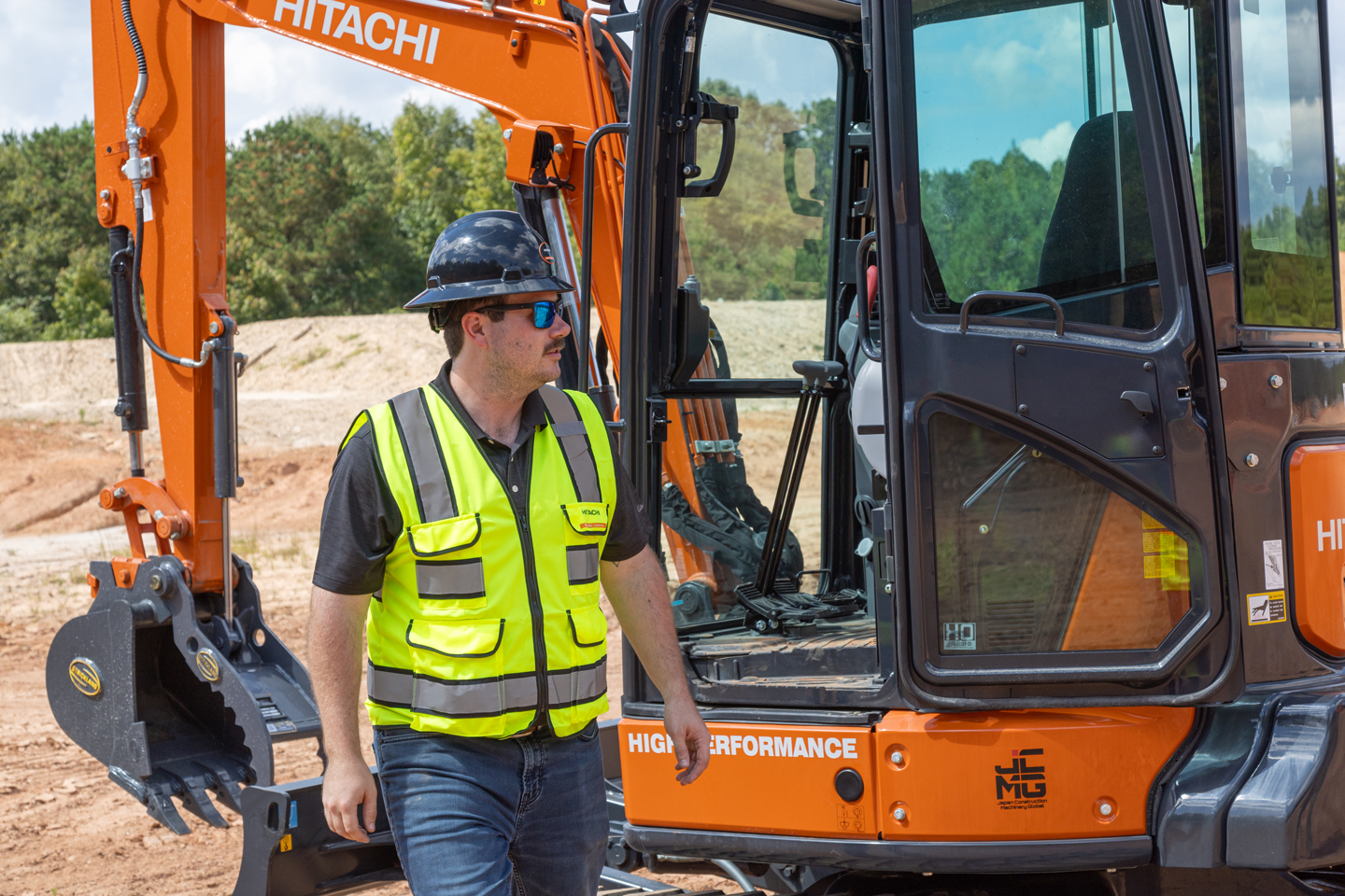 Worker walking next to a Hitachi compact tractor wearing a hi-vis vest and hard hat.