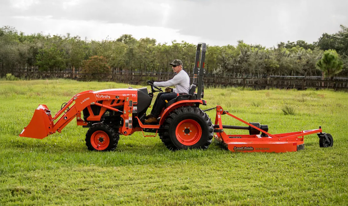 Person on a Kubota tractor with a Land Pride attachment in a green field with a treeline in the background.