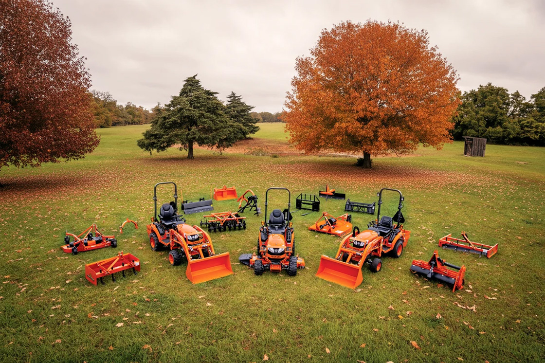 Various Kubota tractors and other pieces of equipment out on a field surrounded by autumn foliage.