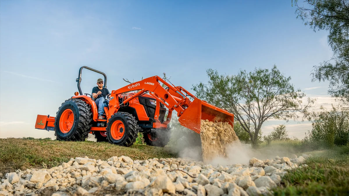 Man in a Kubota tractor dumping gravel onto a rural road near some trees.