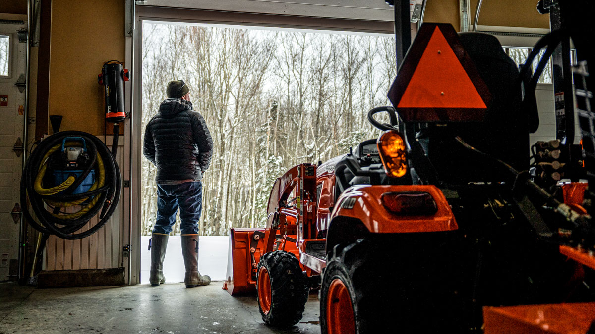 Person standing next to a Kubota tractor in a garage looking out at a winter landscape.